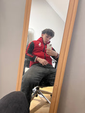 Fiatal férfi tükörszelfi piros Ralph Lauren streetwear mellényben, farmernadrágban, modern enteriőrben.