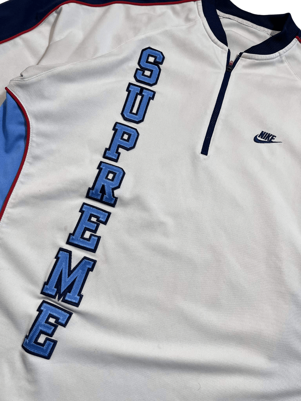 Fehér Nike streetwear felső, SUPREME felirattal, kontrasztos kék-piros részletekkel.